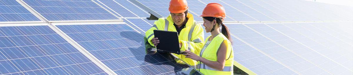 Two people in yellow hi-vis vests and orange safety helmets leaning on a solar panel in a field, looking at an iPad